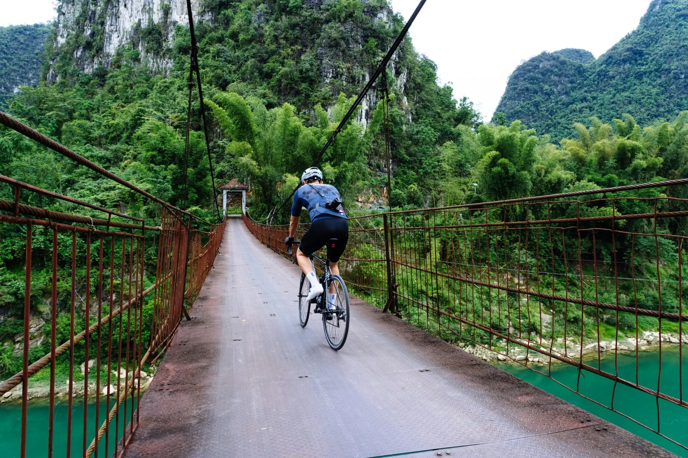 A road cyclist crosses a long suspension bridge framed by limestone karst peaks and tropical forest. The image evokes resilience, journey, and low-carbon exploration.